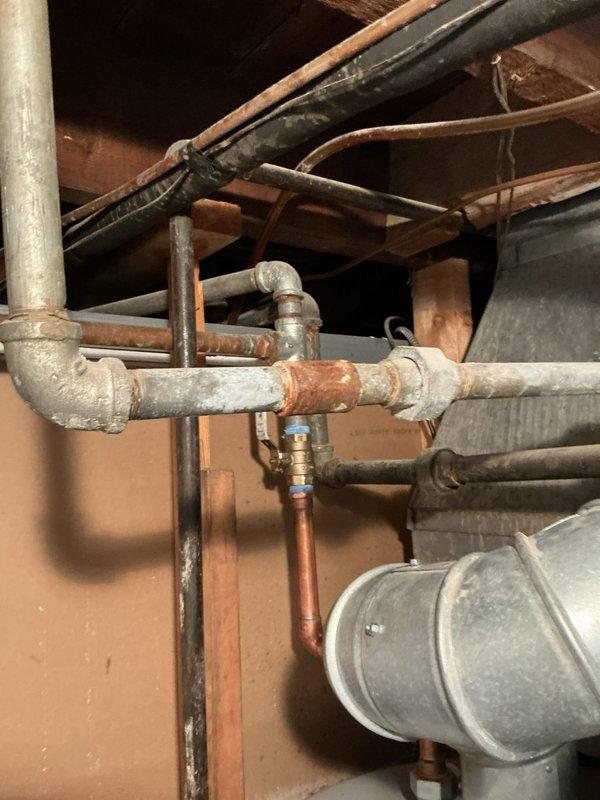 Responded to complaint of no water flow from shower with issue progressively worsening over one year period. Accessed plumbing infrastructure through exposed wall cavity to investigate potential faucet malfunction versus hot water supply problem. Inspected galvanized, copper, and black iron piping systems running vertically and horizontally through basement utility space. Identified significant corrosion on gray horizontal supply pipe at valve connection point, with visible rust accumulation compromising water flow. Examined multiple pipe junctions, fittings, and valve assemblies within the exposed plumbing network. Documented condition of aged infrastructure including corroded galvanized piping, copper supply lines, and associated mechanical connections to determine root cause of diminished water pressure affecting shower fixture.