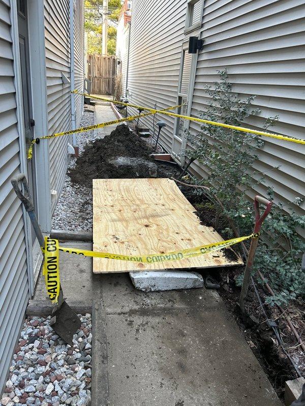 Conducted comprehensive drain and sewer system assessment at residential property requiring urgent attention. Inspection revealed severely deteriorated vertical sewer pipe in excavated access point with extensive corrosion, rust damage, and structural degradation. Standing water with sediment accumulation observed at base of excavation, indicating drainage issues and potential system failure. Corroded metal fittings and connections surrounding main pipe showed advanced deterioration requiring replacement.

Documented conditions in side yard access corridor between structures where active excavation work exposed underground utilities. Area secured with caution tape and plywood covering for safety during assessment. Evidence of recent excavation work visible along concrete walkway with exposed soil and gravel placement. Downspout and drainage components on building exterior evaluated as part of comprehensive system review.

Photographed all critical components including corroded pipe inf