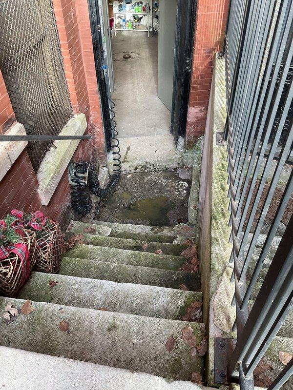 Responded to a water draining issue in the front sewer line of a century-old residence that had been experiencing drainage problems for two days. Upon arrival, inspected the exterior drain grate located in the patio walkway area, which showed significant rust deterioration and potential blockage concerns. The rusted metal grate was embedded in heavily cracked concrete with visible structural deterioration around the drain opening.

Conducted a thorough assessment of the drainage system, including inspection of the lower-level exterior stairway access point where standing water and moisture accumulation were evident on the concrete landing. The presence of moss growth on the stairs and standing water at the base indicated chronic drainage issues and improper water flow from the upper drain system.

Utilized drain cleaning equipment to clear the blockage in the front sewer line, removing accumulated debris and sediment that had built up over time in the aging cast iron or clay pipe syste