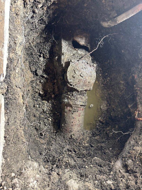 Completed commercial main line sewer installation requiring interior and exterior excavation work. Severely corroded existing drainage system with deteriorated metal components was identified and documented, showing extensive rust buildup and mineral deposits in the concrete channel with standing water. Containment barriers with protective plastic sheeting and dust control measures were installed to isolate the work area from occupied spaces during the excavation and installation process.