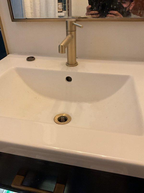 Responded to a slow-draining bathroom sink issue in a century-old home following a referral from another plumbing company. Upon arrival, performed a comprehensive inspection of the bathroom vanity plumbing system, including the modern white ceramic sink with brushed gold fixtures and contemporary P-trap configuration. Examined the drain assembly, overflow mechanism, and verified proper venting. Inspected under-sink cabinet area to assess drain line connections and identify potential blockages or structural issues common in older homes. Documented existing conditions with photographs showing the vanity setup and under-sink plumbing components. Provided customer with findings and recommendations for resolving the drainage concern.