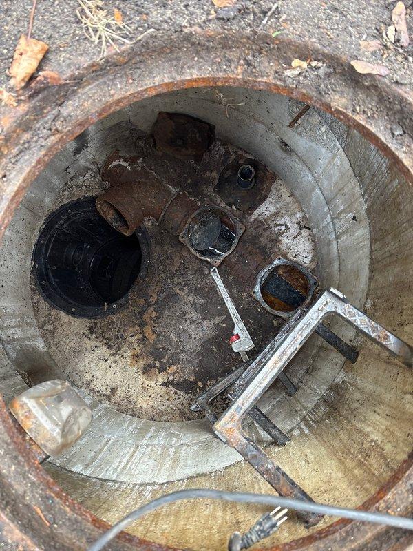 Performed inspection of underground utility access pit and drainage system showing significant corrosion and rust on metal components including valves and connectors. Assessed condition of concrete vault with visible deterioration and rust staining throughout interior walls and rim. Cleared debris and evaluated standing water accumulation in circular drainage opening with corroded metal rim, utilizing hand tools to examine blockage and drainage flow issues requiring repair work and future PDP inspection.