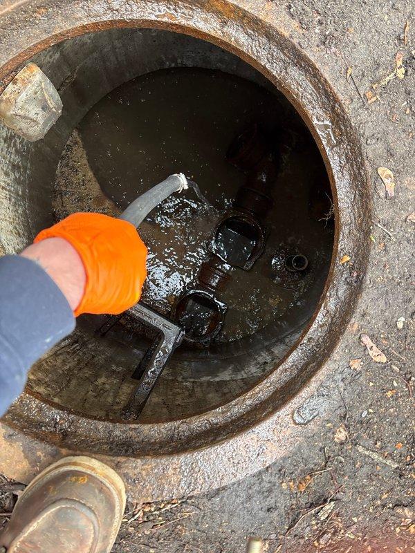 Performed inspection of underground utility access pit and drainage system showing significant corrosion and rust on metal components including valves and connectors. Assessed condition of concrete vault with visible deterioration and rust staining throughout interior walls and rim. Cleared debris and evaluated standing water accumulation in circular drainage opening with corroded metal rim, utilizing hand tools to examine blockage and drainage flow issues requiring repair work and future PDP inspection.
