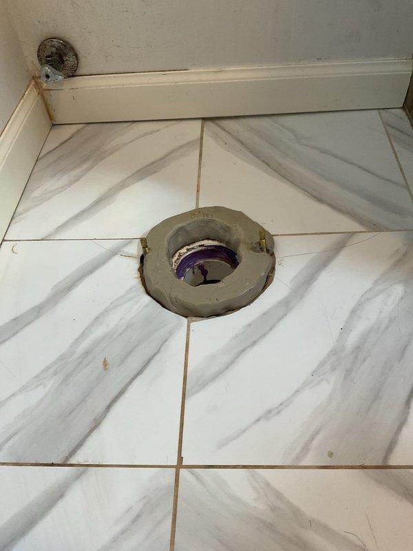 Responded to warranty callback for toilet leaking from base. Upon inspection, found severely corroded toilet flange with visible deterioration and staining around the perimeter. Documented existing conditions showing compromised flange integrity and potential seal failure between toilet base and drain connection. Assessed marble-pattern tile flooring and surrounding area for water damage. Identified purple PVC drain piping beneath flange opening requiring attention to resolve ongoing leak issue at toilet base connection.