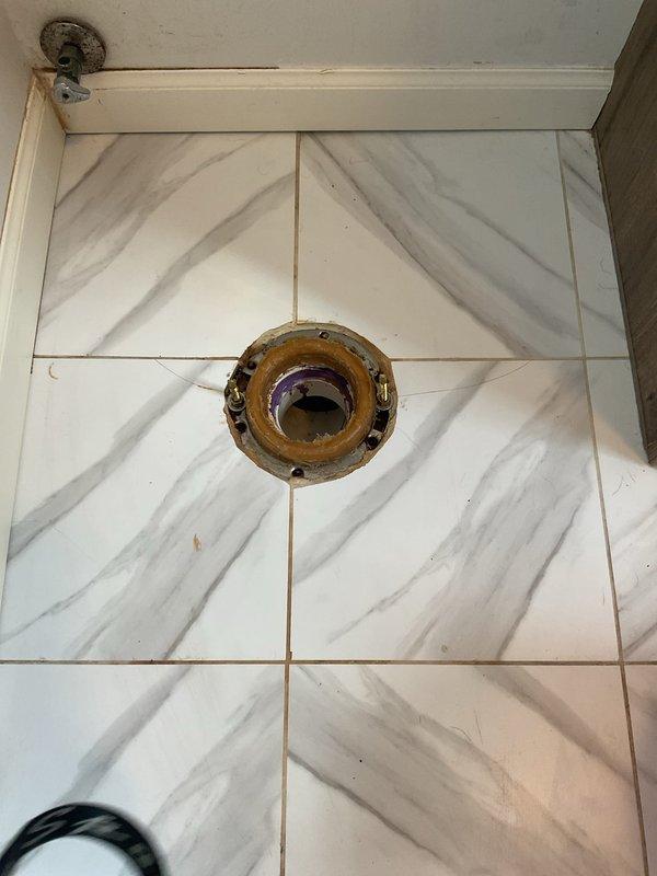 Responded to warranty callback for toilet leaking from base. Upon inspection, found severely corroded toilet flange with visible deterioration and staining around the perimeter. Documented existing conditions showing compromised flange integrity and potential seal failure between toilet base and drain connection. Assessed marble-pattern tile flooring and surrounding area for water damage. Identified purple PVC drain piping beneath flange opening requiring attention to resolve ongoing leak issue at toilet base connection.