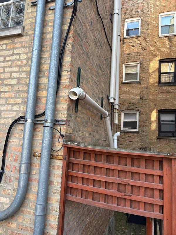Completed inspection of exterior plumbing installation on multi-story brick building, including vertical drainage system and water heater venting. Documented existing PVC pipe configuration with mounting brackets and elbow joints along building exterior. Part ordered and available in shop for scheduled follow-up installation to complete the tanked water heater service.