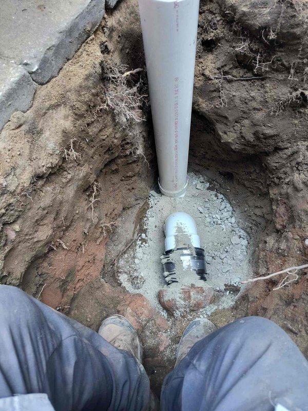 Excavated and exposed deteriorated underground plumbing infrastructure, revealing severely corroded pipe fittings and connections requiring replacement. Installed new white PVC piping system with proper coupling and metal adapter connections, bedding the installation in appropriate sand and gravel material to ensure stable positioning and proper drainage within the trench.