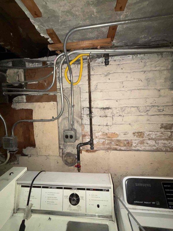 Inspected basement plumbing and utility infrastructure in laundry area with exposed joists and aged brick wall. Documented existing conditions of water supply lines, gas piping with red valve handles, and corroded vertical pipe requiring future attention. Photographed gray electrical conduit, flexible yellow cable routing, and overall system layout for customer records and potential repair planning.