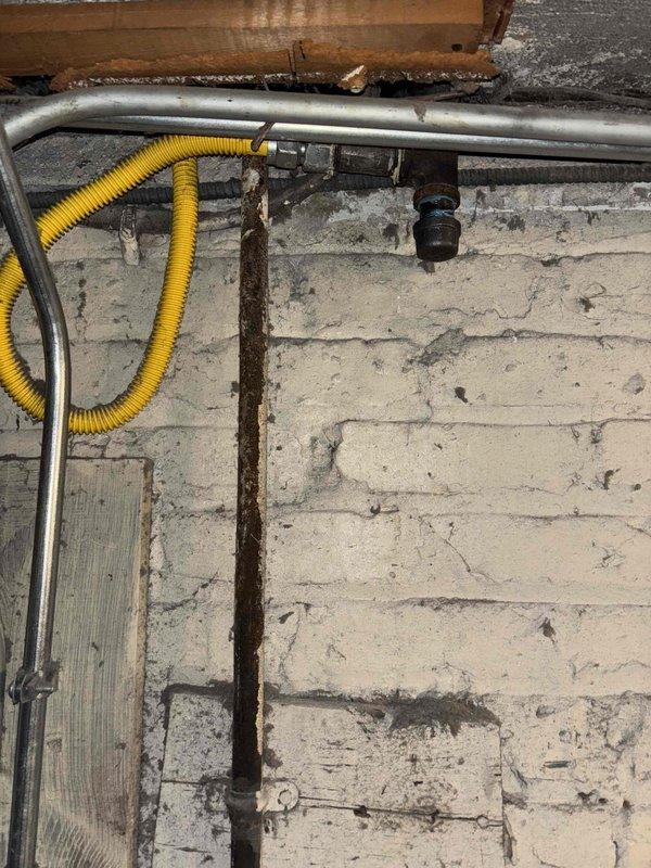 Inspected basement plumbing and utility infrastructure in laundry area with exposed joists and aged brick wall. Documented existing conditions of water supply lines, gas piping with red valve handles, and corroded vertical pipe requiring future attention. Photographed gray electrical conduit, flexible yellow cable routing, and overall system layout for customer records and potential repair planning.
