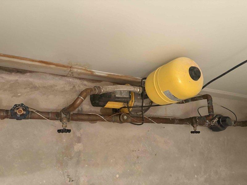 Responded to customer complaint of low water pressure throughout entire building. Upon investigation, inspected the overhead-mounted hydronic system including expansion tank, circulator pump, and associated piping network. Examined the yellow expansion tank with pressure valve, circulation pump unit, and copper distribution piping showing signs of oxidation and age-related discoloration. Assessed all visible components including shutoff valves, flexible connectors, and mounting hardware. Photographed system configuration for documentation purposes. Identified potential issues with the pressurization system that could be affecting water delivery throughout the building. System components appeared functional but showed normal operational wear consistent with age of installation.