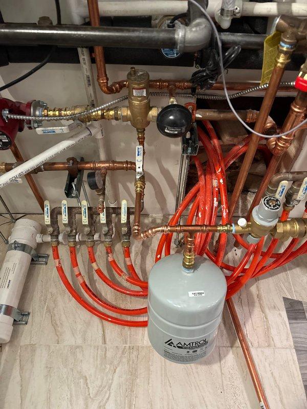Installed comprehensive hydronic heating distribution manifold system in mechanical room. System includes multiple zone configuration with red PEX tubing lines, copper piping with brass fittings, and series of individual ball valves for zone control. Mounted Amtrol expansion tank and integrated pressure control components. All manifold connections secured with proper brass fittings, zone identification tags applied to valves for future reference. System tested and operational for multi-zone radiant heating distribution.