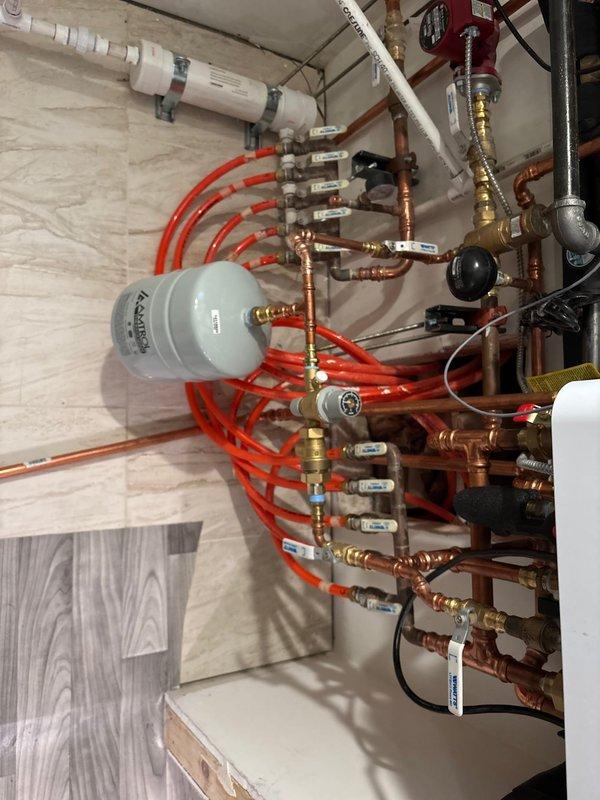 Installed comprehensive hydronic heating distribution manifold system in mechanical room. System includes multiple zone configuration with red PEX tubing lines, copper piping with brass fittings, and series of individual ball valves for zone control. Mounted Amtrol expansion tank and integrated pressure control components. All manifold connections secured with proper brass fittings, zone identification tags applied to valves for future reference. System tested and operational for multi-zone radiant heating distribution.