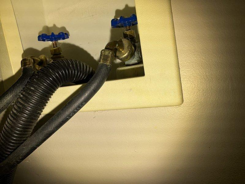 Responded to service request for corroded laundry water faucet with stuck handle that would not turn off. Upon arrival, inspected recessed wall box containing water supply valves with blue cross-handles and connected black corrugated washing machine supply line. Assessed condition of brass valve bodies showing age and patina, and evaluated both upper and lower shut-off valves for functionality and corrosion damage. Provided quote for repair or replacement options to address the inoperable valve handle and ensure proper water flow control to the laundry appliance.