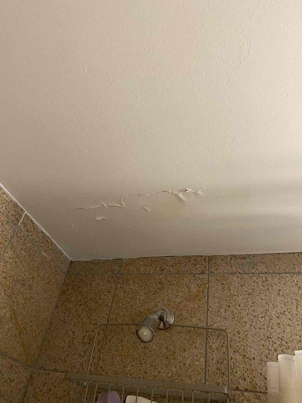 Responded to water leak complaint originating from upper unit bathroom and affecting the unit below. Upon arrival, inspected the bathroom and identified visible ceiling damage with peeling paint and surface deterioration in the lower unit, indicating active water intrusion. Examined the shower area in the upper unit, focusing on the tile surround, grout lines, shower fixtures, and wall penetrations. Documented water-damaged ceiling showing multiple areas of paint failure and underlying material exposure. Assessed shower enclosure with beige marble-pattern tile, chrome fixtures including rain showerhead, grab bar, and mixing valve for potential leak sources. Photographed evidence of damage for documentation purposes and to establish scope of issue for necessary repairs.