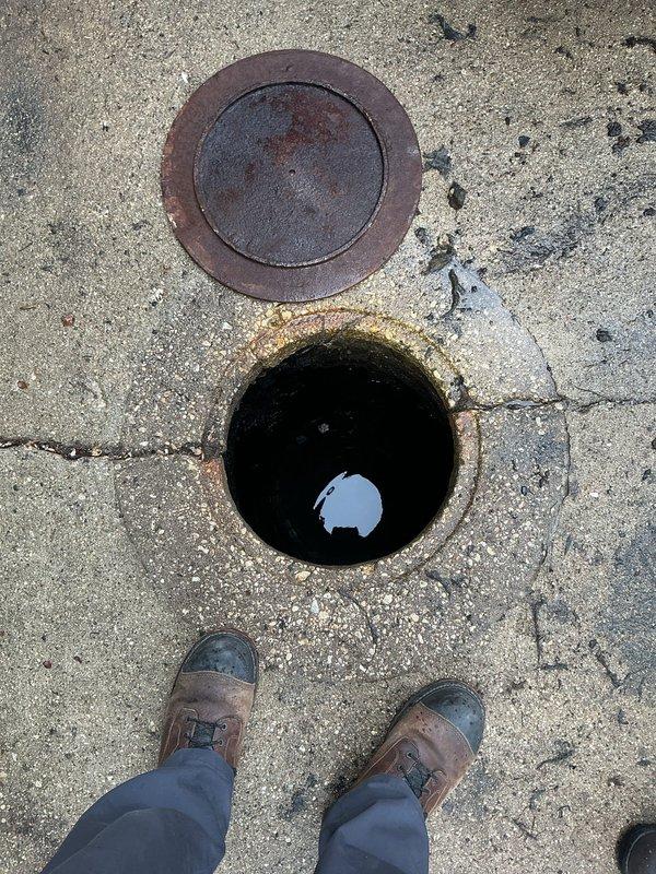 Completed follow-up inspection of underground utility access structure. Documented current condition of manhole with two access points, noting significant deterioration of interior concrete walls with extensive staining, mineral deposits, and biological growth. Photographed standing water at bottom of structure showing contamination and debris accumulation. Observed rust and corrosion around rim edges of uncovered opening, with weathered metal cover present on adjacent access point.