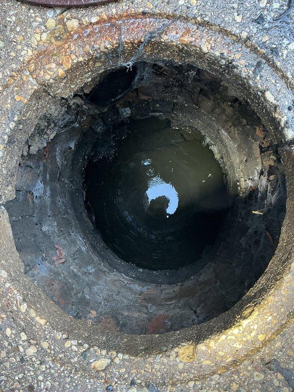 Completed follow-up inspection of underground utility access structure. Documented current condition of manhole with two access points, noting significant deterioration of interior concrete walls with extensive staining, mineral deposits, and biological growth. Photographed standing water at bottom of structure showing contamination and debris accumulation. Observed rust and corrosion around rim edges of uncovered opening, with weathered metal cover present on adjacent access point.