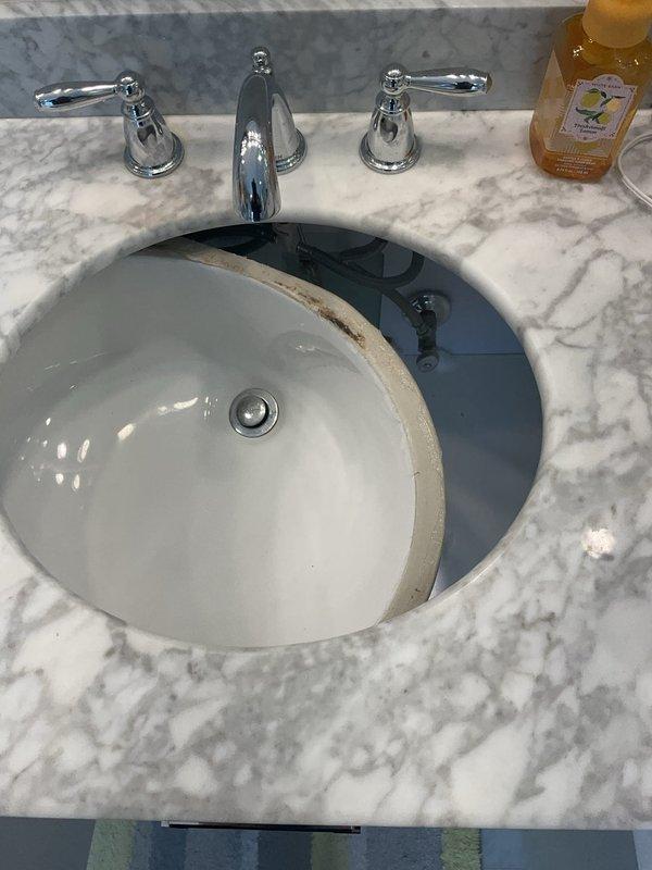 Assessed bathroom vanity sink installation with undermount white porcelain basin set into white marble countertop with gray veining. Identified rust-colored staining along sink rim where basin meets countertop, indicating potential water damage or seal failure. Evaluated existing three-piece polished chrome faucet assembly with center spout and dual lever handles, all in functional condition. Documented condition of undermount installation and chrome pop-up drain stopper assembly for replacement estimate. Provided assessment for complete vanity replacement due to compromised countertop integrity at sink mounting area.
