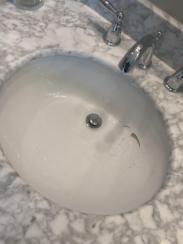 Assessed bathroom vanity sink installation with undermount white porcelain basin set into white marble countertop with gray veining. Identified rust-colored staining along sink rim where basin meets countertop, indicating potential water damage or seal failure. Evaluated existing three-piece polished chrome faucet assembly with center spout and dual lever handles, all in functional condition. Documented condition of undermount installation and chrome pop-up drain stopper assembly for replacement estimate. Provided assessment for complete vanity replacement due to compromised countertop integrity at sink mounting area.