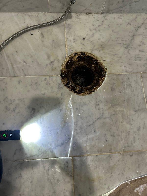 Responded to service call for floor drain inspection and maintenance. Upon arrival, found heavily corroded and deteriorated floor drain assembly in marble-tiled shower area with significant rust staining and oxidation around the drain flange and interior components. Removed existing drain cover and strainer assembly to assess condition of drain body and surrounding fixture. Documented extent of corrosion damage affecting the metal drain components. Evaluated drain opening integrity and water flow capacity to determine if replacement parts or complete drain assembly replacement would be required to restore proper function and prevent future leaks or drainage issues.