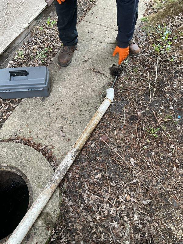 Responded to clogged drain service call and performed rodding operation from catch basin to main sewer line. Accessed the outdoor drainage system through concrete-mounted manhole opening, which showed standing water and significant corrosion on metal components. Utilized specialized drain cleaning equipment and long-handled rodding tools to clear blockage between catch basin and sewer connection. Customer advised that camera inspection video will be required to assess line condition and identify any underlying issues with the drainage system.