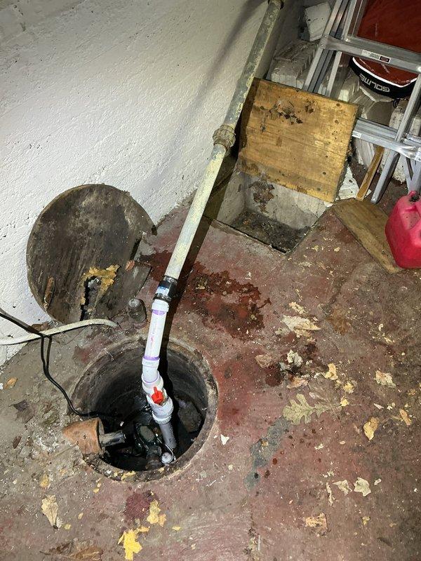 Completed callback to inspect and photograph existing sump pump system and deteriorated flooring conditions. Documented corroded floor drain assembly with exposed subfloor damage and significant water staining on concrete basement floor surrounding sump pit. Observed functional sump pump installation with PVC discharge piping and check valve in place, though extensive floor deterioration and rust patterns indicate previous water intrusion issues requiring attention.