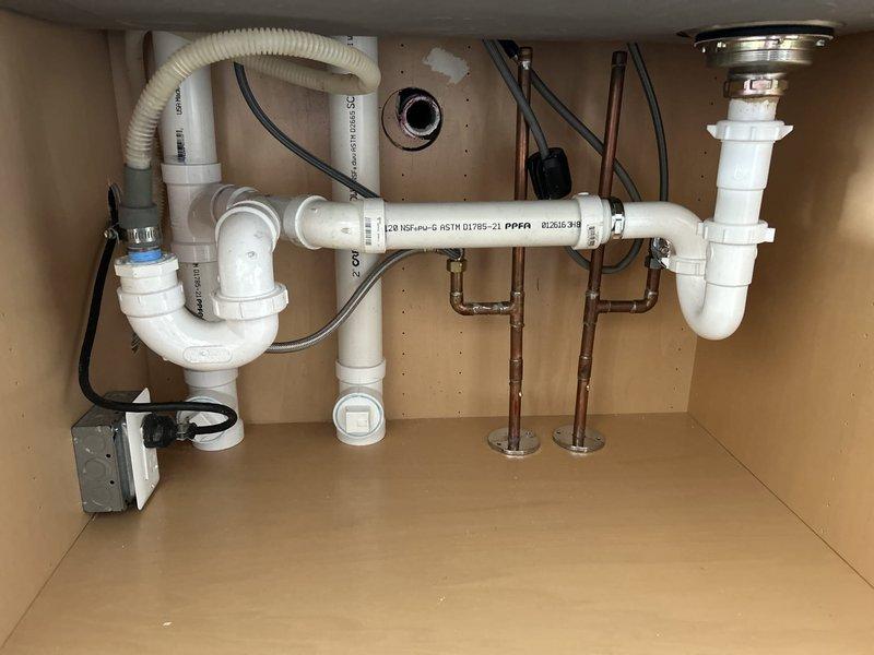 Cored through the base of the kitchen cabinet and installed 2" PVC drain pipework with cleanout access. Ran drain lines into the basement and connected to existing galvanized drain and vent system using proper fastening hangers and support brackets. Installed dedicated P-trap for dishwasher discharge line and separate P-trap assembly for right side basin. Connected garbage disposal unit with corrugated flexible discharge hose to drain system. Installed water supply lines using braided stainless steel flex lines connected to chrome shutoff valves and copper supply piping. Mounted water filtration device horizontally within cabinet space and integrated into supply system. All connections tested and verified for proper drainage and no leaks.