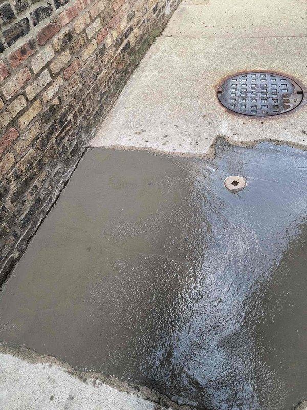 Assessed exterior drainage issue in alleyway between buildings where significant water pooling was observed on concrete surface near brick structures. Identified failed or inadequate drainage system causing water accumulation despite presence of existing drain infrastructure. Area was cordoned off with caution tape for safety due to standing water hazard. Documented conditions showing water collection on ground surface with nearby circular drain cover that was not effectively managing water flow. Repair has been sold and awaiting parts arrival to complete drainage system repair work.
