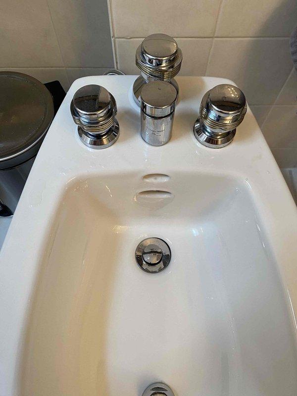 Responded to service call for leaking bidet fixture. Upon inspection, identified leak originating from supply line connection beneath the bidet. Tightened braided flexible supply line connection at the threaded fitting to eliminate water seepage. Verified all chrome-plated faucet components and pop-up drain assembly were functioning properly with no additional leaks detected. Documented fixture condition with photos showing three-handle bidet configuration and underlying plumbing connections for service records.