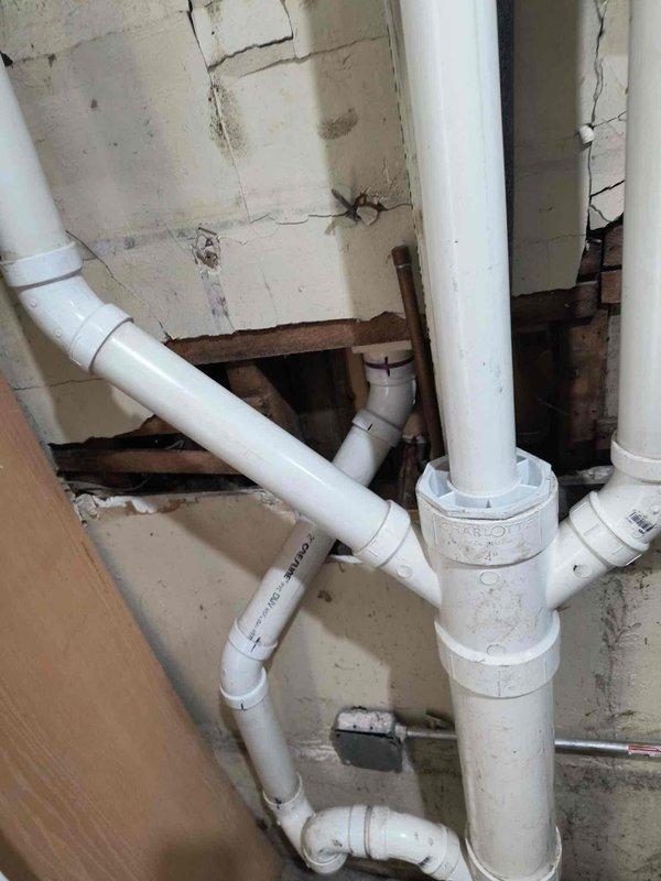 Completed plumbing inspection and documentation of basement drainage system featuring white PVC piping with multiple joints, elbows, and vertical drain lines. Identified significant water accumulation on concrete floor indicating active drainage or seepage issues requiring follow-up repair work. Documented deteriorated wall conditions with structural cracks and aging plumbing infrastructure for repair scheduling and scope determination.