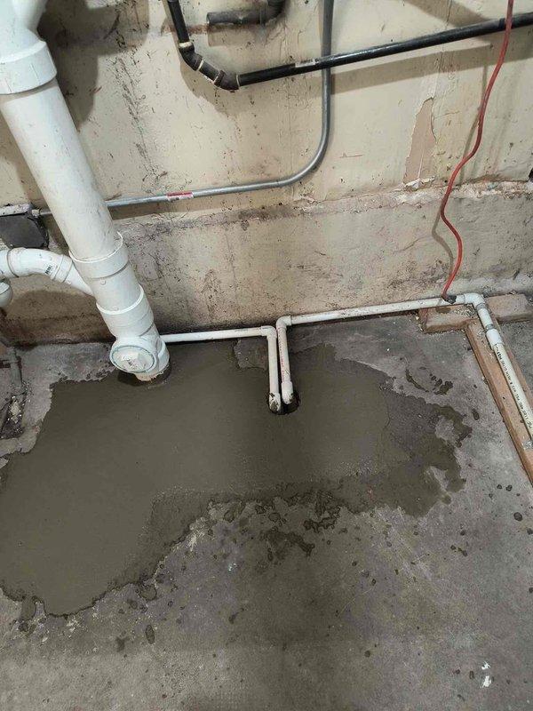 Completed plumbing inspection and documentation of basement drainage system featuring white PVC piping with multiple joints, elbows, and vertical drain lines. Identified significant water accumulation on concrete floor indicating active drainage or seepage issues requiring follow-up repair work. Documented deteriorated wall conditions with structural cracks and aging plumbing infrastructure for repair scheduling and scope determination.