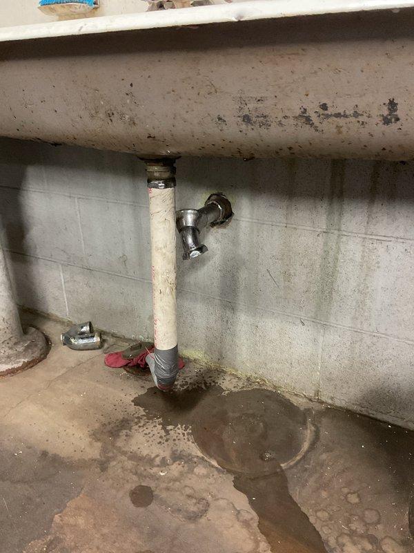 Responded to commercial plumbing service call for completely clogged sink with no drainage. Upon arrival, accessed the drain system beneath the fixture and inspected the vertical drain pipe, P-trap, and connection points. Cleared the obstruction from the drain line, restoring full drainage function. Inspected the plumbing assembly including the chrome P-trap fitting, vertical drain pipe, and floor connection boot for proper operation and potential leaks. Verified all connections were secure and tested the system to confirm water was flowing freely through the drain without backup or slow drainage issues.