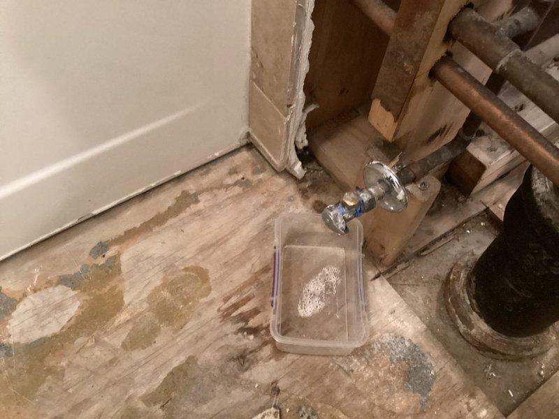 Responded to priority call for leaking toilet valve. Upon arrival, found chrome-plated shut-off valve actively leaking at the connection point in an unfinished utility area with exposed floor joists and rough plumbing. Placed collection container beneath the valve to contain water discharge and prevent further damage to subflooring. Assessed the condition of the copper supply line and valve assembly, noting signs of age and corrosion on surrounding plumbing components. Documented existing conditions including stained subflooring and exposed wall framing for reference. Valve required immediate attention to stop active leak and restore proper water control to the toilet fixture.