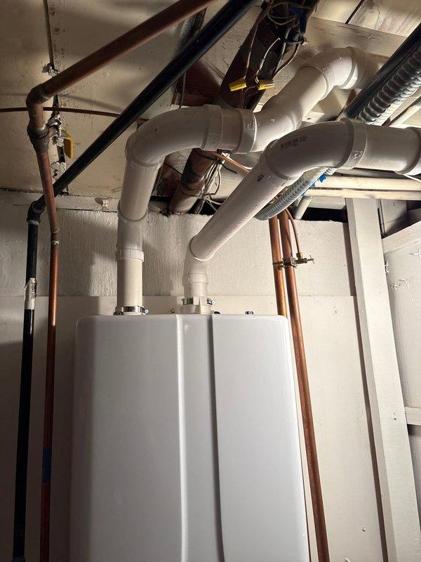 Completed tankless water heater installation in mechanical room, mounting dual units side by side on wall with proper clearances. Installed white PVC venting system with appropriate elbows and connections, routing exhaust pipes through ceiling infrastructure and terminating through exterior stone wall. Reworked return line using 1/2" copper piping, incorporating boiler drain and check valve as specified. Connected copper water supply lines vertically along units and integrated with existing ceiling plumbing network. Configured exterior PVC venting with multiple 90-degree elbows to navigate around building features and ensure proper termination clearance from structure. All connections secured and tested for proper operation.