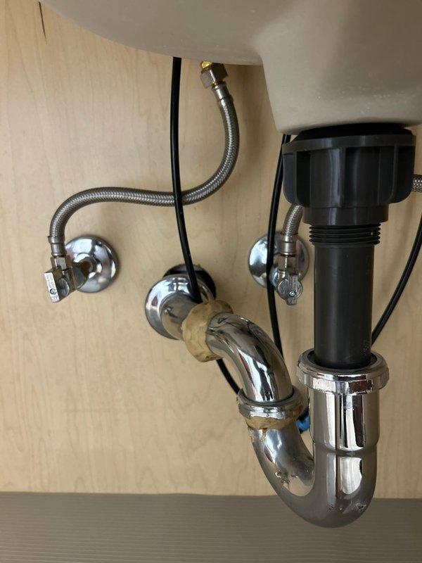 Completed building shut down plumbing repair involving under-sink installation with new chrome P-trap assembly, flexible braided stainless steel supply lines with brass connectors, and wall-mounted shut-off valves with chrome escutcheons. Installed garbage disposal connection components including black plastic discharge fitting with threaded chrome collar, ensuring proper drainage configuration and secure mounting to cabinet backing. All chrome fixtures installed without corrosion, supply lines properly connected to wall-mounted valves, and electrical connections completed with visible power indicator for disposal unit operation.