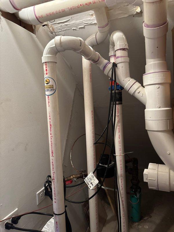 Completed installation of sump pump system with battery backup in basement utility area. Installed comprehensive PVC discharge piping configuration with multiple elbows and vertical runs, secured all electrical connections and cables with proper fastening, and mounted StormCell 10-amp battery backup unit by Liberty Pumps with digital display and multi-function status indicators to ensure continuous operation during power outages.