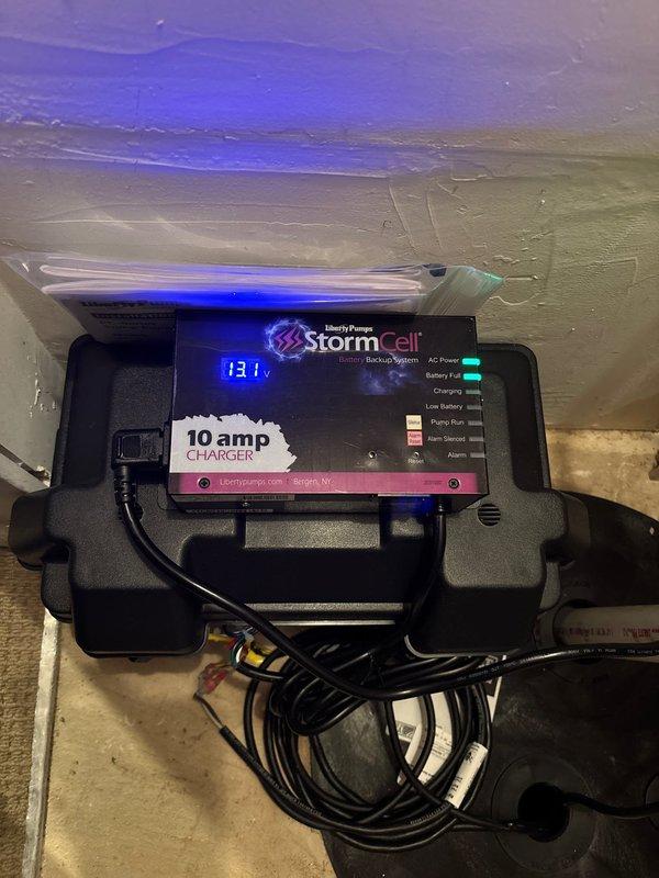 Completed installation of sump pump system with battery backup in basement utility area. Installed comprehensive PVC discharge piping configuration with multiple elbows and vertical runs, secured all electrical connections and cables with proper fastening, and mounted StormCell 10-amp battery backup unit by Liberty Pumps with digital display and multi-function status indicators to ensure continuous operation during power outages.