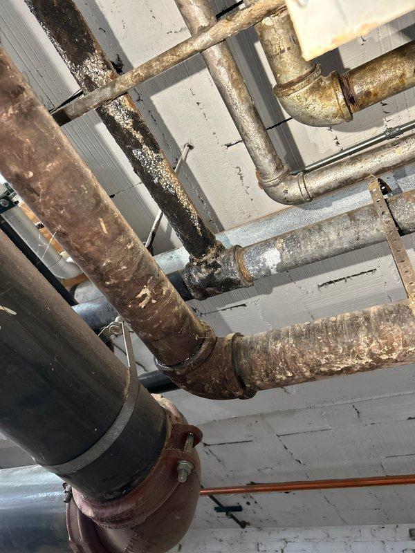 Responded to water leak complaint near laundry room area. Upon inspection, located source of slow leak in basement mechanical space affecting multiple corroded supply lines running along ceiling near laundry area. Extensive rust and oxidation observed throughout exposed piping system, indicating aged infrastructure with compromised integrity. Documented deteriorated pipe conditions showing heavy corrosion on horizontal runs, multiple elbow joints, and connecting fittings secured with metal hangers to ceiling structure.