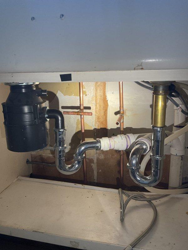 Conducted comprehensive walk-through inspection of commercial property's drain and sewer systems, documenting existing plumbing infrastructure in basement mechanical room and kitchen areas. Photographed complex piping network including copper supply lines, black iron pipes, PVC conduits, and drainage systems; assessed dual-sink kitchen plumbing configuration with garbage disposal unit, chrome P-trap assemblies, and brass drain connections; evaluated condition of exposed plumbing systems, pipe connections, and potential areas of concern including water staining around wall penetrations for estimate preparation.