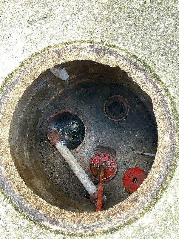 Performed comprehensive inspection and maintenance on residential flood control system located in front yard utility vault. System had not received professional service in several years but was reportedly functioning properly. Accessed underground flood control chamber through concrete vault opening, which showed typical aging with moss and algae growth on surrounding surfaces. 

Inspected all primary components including inlet pipes, discharge valves, and control mechanisms. Examined white PVC inlet conduit with cap fitting for proper sealing and structural integrity. Evaluated two floor drain openings for debris accumulation and proper flow capacity. Assessed red valve assembly and actuator mechanism for operational functionality and signs of wear. Checked metal float arm and linkage system for freedom of movement and correct positioning.

Noted chamber interior showed moderate sediment buildup and staining consistent with age and normal operation. All mechanical components were foun