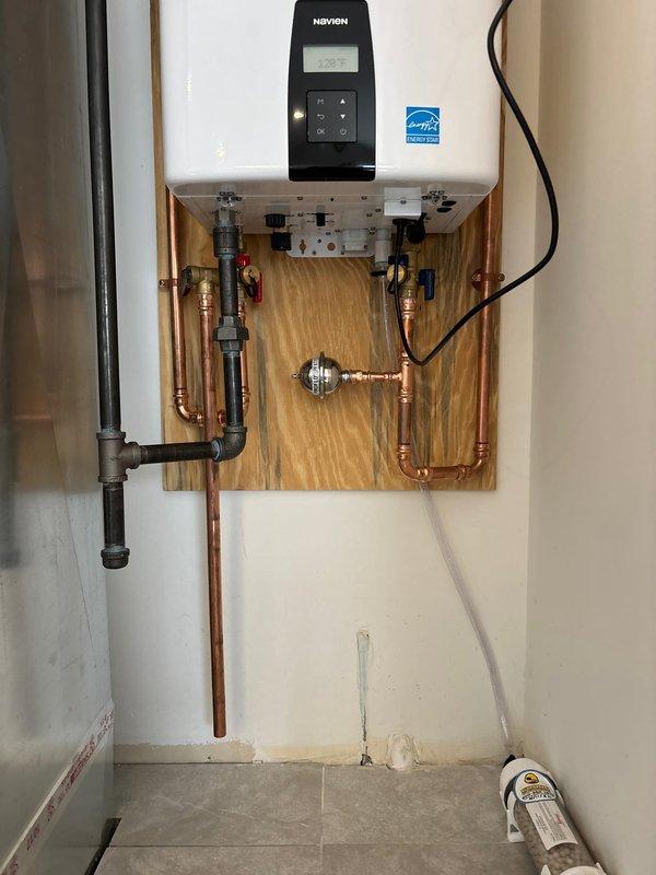 Completed installation of Navien tankless water heater system with full plumbing integration. Mounted unit on wooden backing board with proper structural support and installed complete copper piping manifold including shut-off valves, pressure gauges, and brass fittings on both supply and return lines. Connected gas line with appropriate compression fittings and black pipe. Configured control panel settings and verified proper operation of digital display and Energy Star certified unit. Installation included bathroom plumbing fixtures with chrome shower control valve, tub spout, and drain assembly coordinated with white subway tile surround. All connections tested and verified leak-free with system operational.