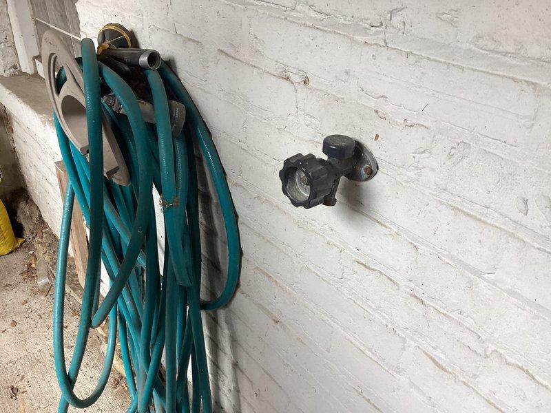 Responded to outdoor spigot backup issue where water was leaking into storage unit at the shut-off valve location. Inspected exterior hose bib mounted on white brick wall and traced supply line to interior copper piping with blue-handled shut-off valve in corner of storage area. Identified leak at valve connection point and resolved water intrusion issue to prevent further damage to storage space.