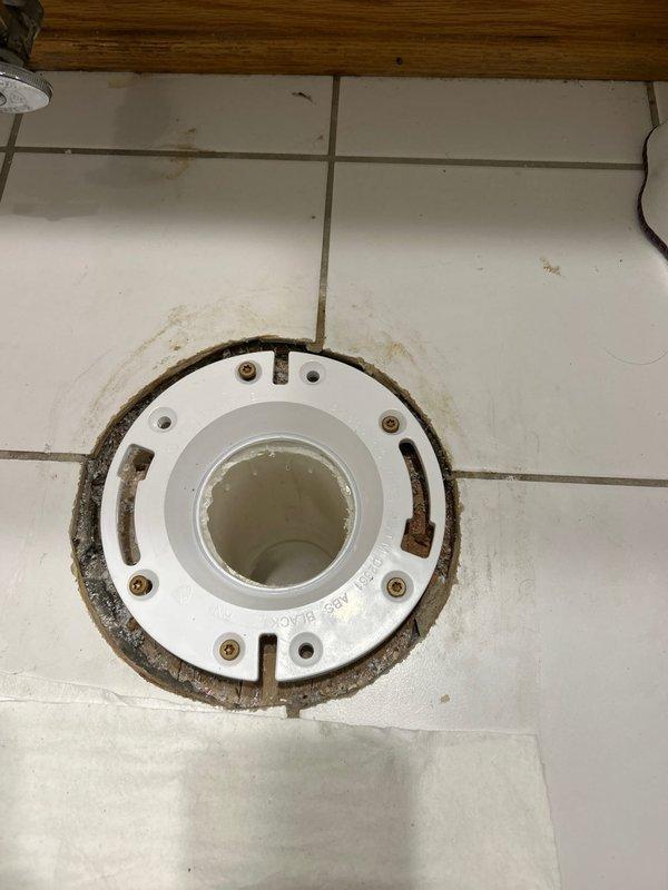Inspected and documented existing floor drain assembly showing significant rust staining and corrosion around the white plastic drain cover and mounting hardware. The drain flange exhibited extensive rust discoloration extending onto surrounding white tile floor, with corroded metal fasteners and water-related mineral deposits indicating long-term moisture exposure and aging fixture components requiring replacement or remediation.
