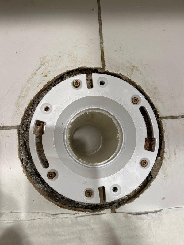 Inspected and documented existing floor drain assembly showing significant rust staining and corrosion around the white plastic drain cover and mounting hardware. The drain flange exhibited extensive rust discoloration extending onto surrounding white tile floor, with corroded metal fasteners and water-related mineral deposits indicating long-term moisture exposure and aging fixture components requiring replacement or remediation.