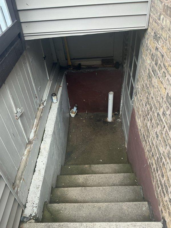 # Flood Control System Installation - Exterior Basement Stairwell

Completed residential drainage assessment and flood control installation for exterior basement stairwell entrance experiencing moisture accumulation and water drainage issues. The concrete stairwell, consisting of 5-6 weathered steps descending between wooden siding and brick walls, showed evidence of persistent water pooling at the landing area with debris accumulation and moisture damage.

Evaluated existing drainage infrastructure including white PVC pipe at landing and assessed water flow patterns contributing to flooding concerns. Documented deteriorated concrete surfaces and inadequate drainage capacity at the lower level entrance point. Recommended and prepared site for flood control system installation to redirect water away from basement access point and prevent future water intrusion. Inspected crawlspace ventilation access panels and surrounding grade conditions to ensure comprehensive drainage solution addre