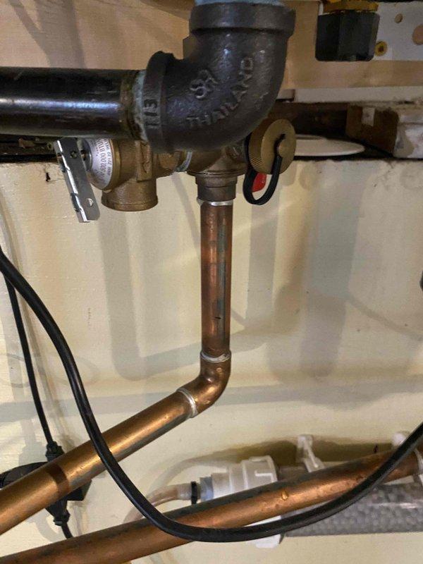 Responded to warranty service call for plumbing system inspection in mechanical space. Upon arrival, evaluated pressure relief valve assembly and associated piping connections mounted beneath floor joists, including cast iron elbow fitting with brass valves and copper piping configuration. Documented existing plumbing infrastructure showing aged copper lines with sensor wiring secured along pipe runs, connected to water heating appliance. Captured photographic documentation of installation for warranty assessment and coordination with management team for appropriate technician assignment and follow-up action.