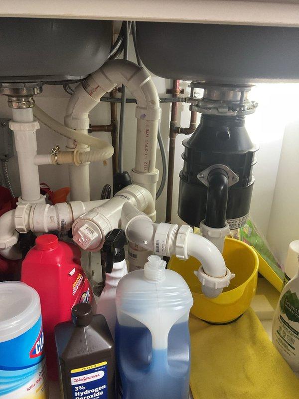 Responded to service call for water leaking from garbage disposal unit. Upon inspection, identified leak source at disposal mounting assembly and drain connections beneath double-basin kitchen sink. Tightened mounting bolts and discharge elbow connection, resealed drain flange, and verified all PVC drain line connections for proper seal. Placed collection bucket beneath unit to monitor for residual dripping. Tested system operation and confirmed leak was successfully repaired with no further water discharge observed.