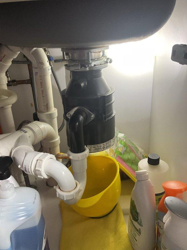 Responded to service call for water leaking from garbage disposal unit. Upon inspection, identified leak source at disposal mounting assembly and drain connections beneath double-basin kitchen sink. Tightened mounting bolts and discharge elbow connection, resealed drain flange, and verified all PVC drain line connections for proper seal. Placed collection bucket beneath unit to monitor for residual dripping. Tested system operation and confirmed leak was successfully repaired with no further water discharge observed.