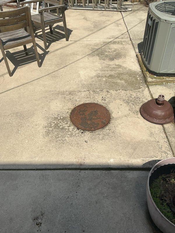 Conducted comprehensive site assessment for residential sewer repair estimate on outdoor patio area. Inspected corroded circular metal sewer access cover showing significant rust deterioration, evaluated concrete pad condition with visible weathering and surface staining, and assessed accessibility around HVAC condenser units and landscape features. Documented existing conditions through photographic survey to determine scope of work required for sewer line repair and access point restoration.