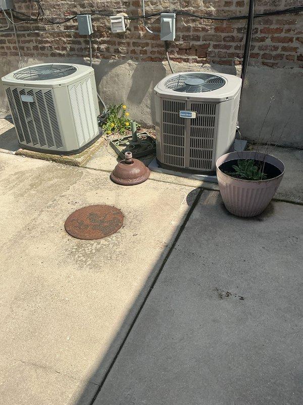 Conducted comprehensive site assessment for residential sewer repair estimate on outdoor patio area. Inspected corroded circular metal sewer access cover showing significant rust deterioration, evaluated concrete pad condition with visible weathering and surface staining, and assessed accessibility around HVAC condenser units and landscape features. Documented existing conditions through photographic survey to determine scope of work required for sewer line repair and access point restoration.