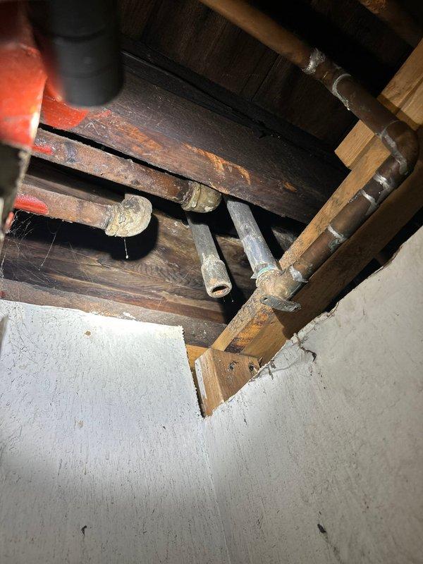 Responded to emergency call for ceiling pipe leak in basement originating from utility room area near riser. Accessed ceiling cavity through existing opening and inspected aged metal piping system running horizontally through wooden framing structure. Identified leak source at pipe joint connection near riser location. Documented conditions with photographs showing oxidized piping, structural framing, and affected areas for service records.